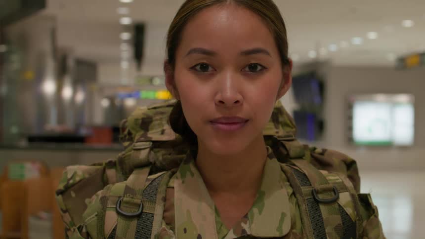 A female soldier in full military uniform carrying a backpack stands tall in a portrait taken at an airport