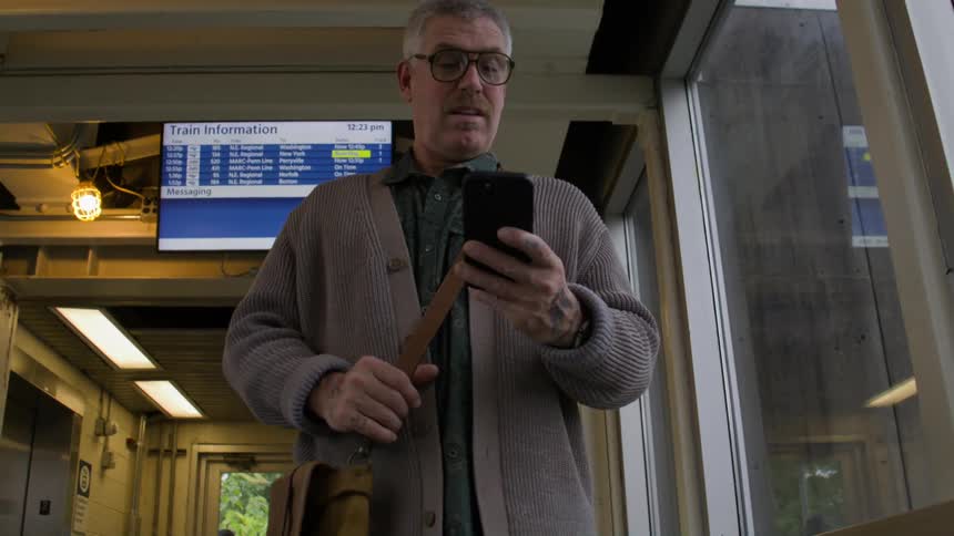 A man in a train station looks up at a digital screen and then down at his phone