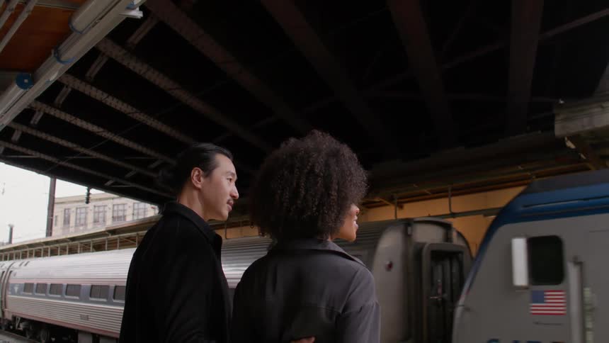 A young couple waits at the train station as a train arrives on the tracks