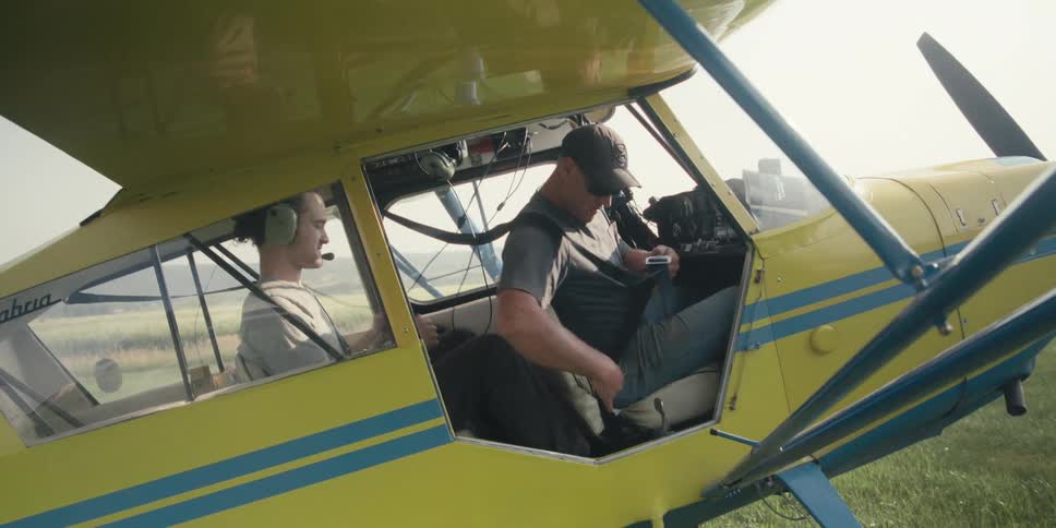 Two men are inside a yellow and blue small airplane with the pilot buckling in