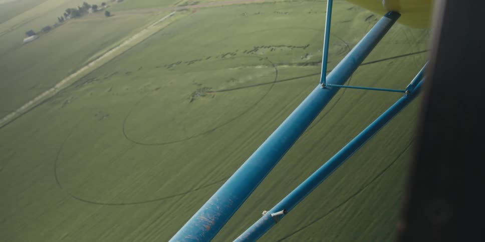 Man piloting a small aircraft over farmland looks toward a field of green grain