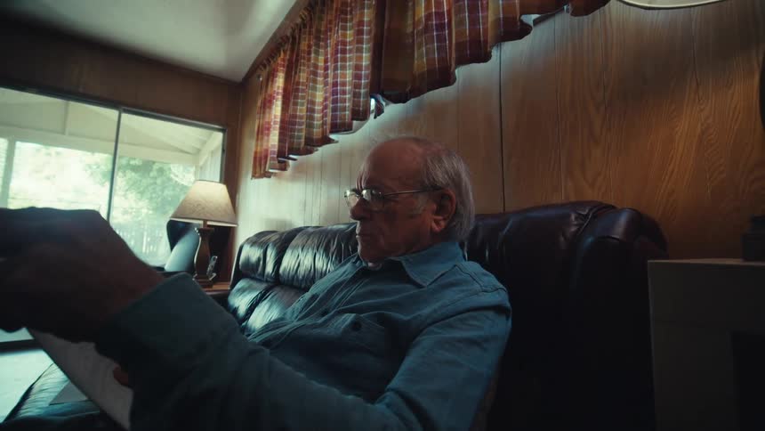An elderly man is seated on a leather couch reading in a woodpaneled room