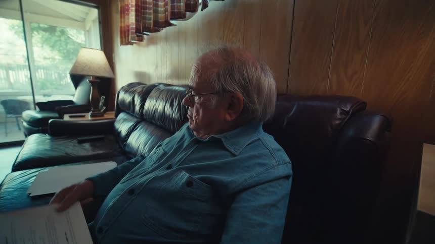 An elderly man is sitting on a couch at home reading documents