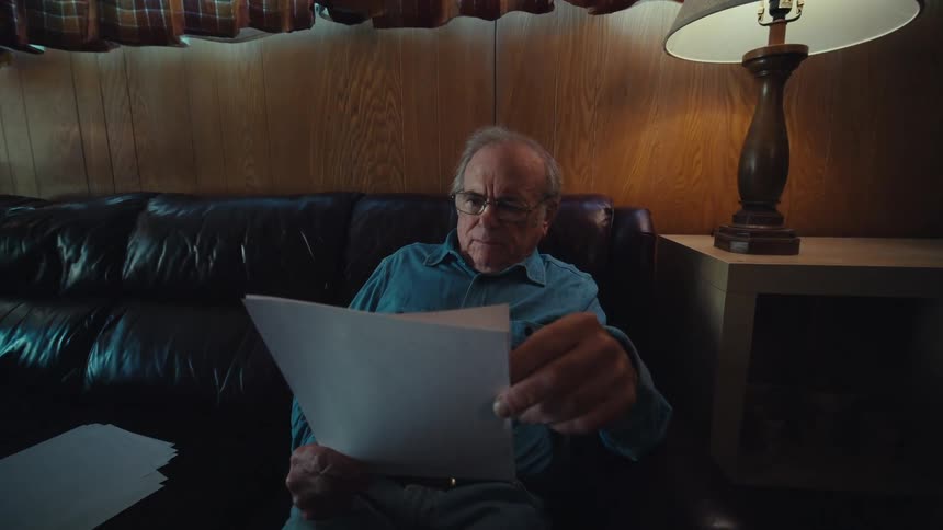 An elderly man sitting on a couch at home reading documents