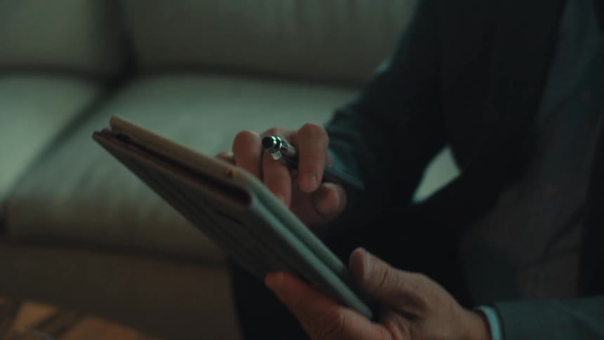 A man in a suit is sitting on a white sofa and fiddling with a tablet and he has a pen between some of his fingers