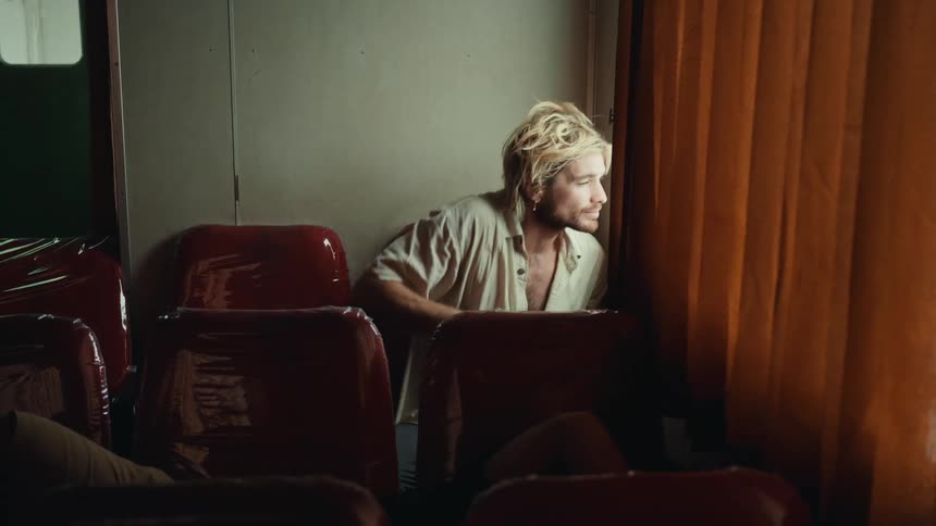 A blond man is sitting on a red seat and is squinting