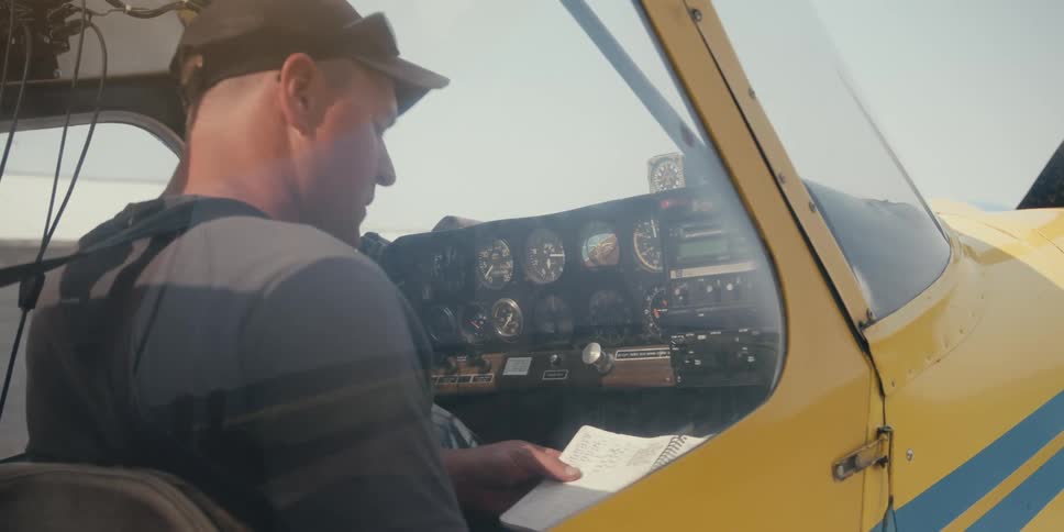 A person is seated inside the cockpit of a small yellow aircraft taking notes in front of the instrument panel and controls