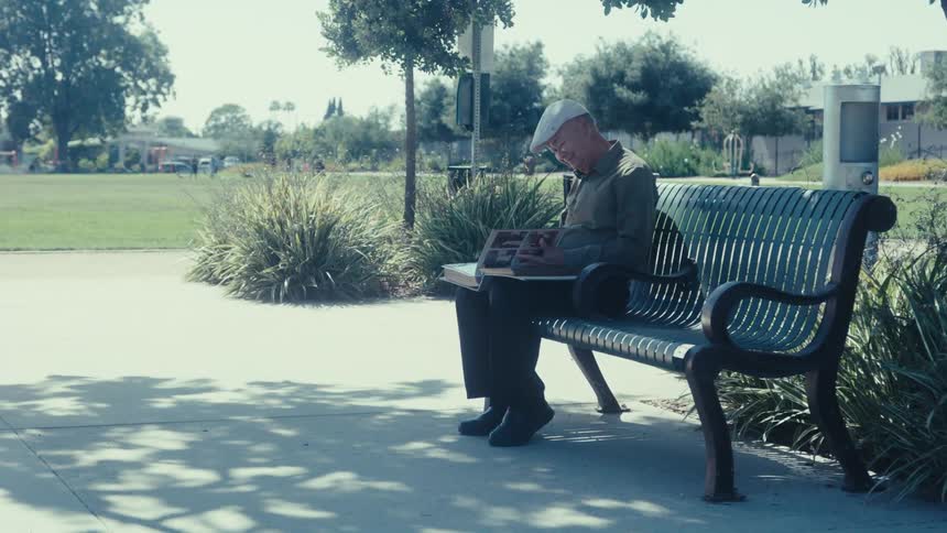An elderly man sitting on a park bench looking at a photo album
