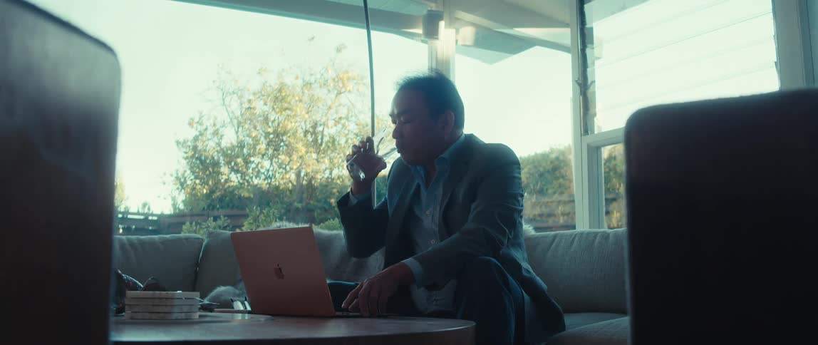 A person in a suit sits on a couch in a welllit room using a laptop on a coffee table while drinking water