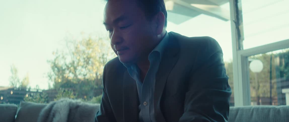 A man in a suit is sitting on a couch in a well lit room working intently on his laptop placed on a coffee table in front of him
