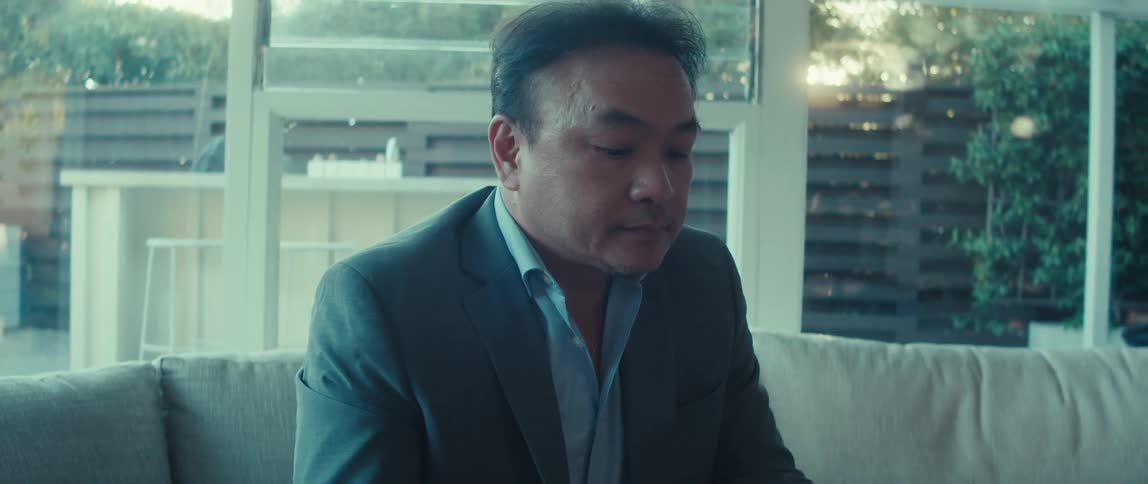 A man in a business suit sits on a couch at home using a laptop on a sunny morning