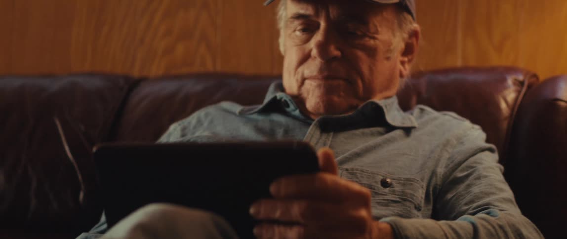An elderly man wearing a cap and a blue shirt sits on a brown leather couch using a tablet in the room