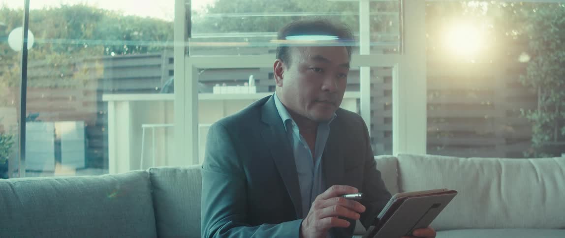 A man in a business suit sits on a couch at home using a tablet on a sunny morning