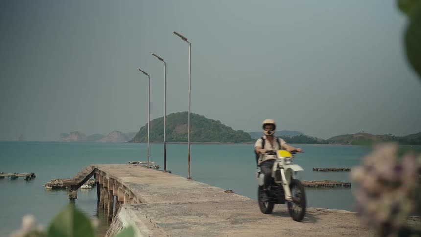 A man wearing a white helmet a shirt and a black backpack rides a dirt bike off a pier with a serene sea in the background