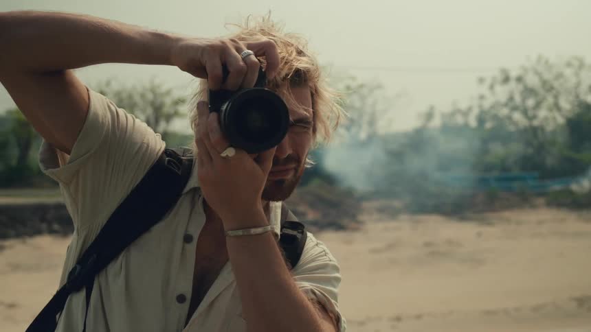 A person wearing a white shirt and a backpack is sitting on a motorcycle holding a camera and taking a photograph