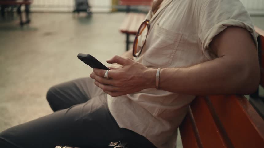 A blonde man sits on a bench in a coastal park using his phone