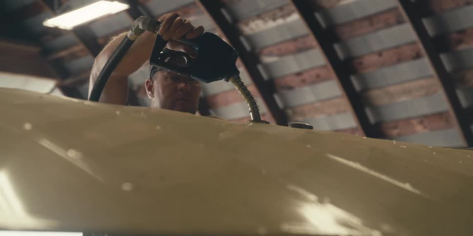 A person is refueling an aircraft inside a hangar with the fuel nozzle inserted into the aircraft wing fuel tank