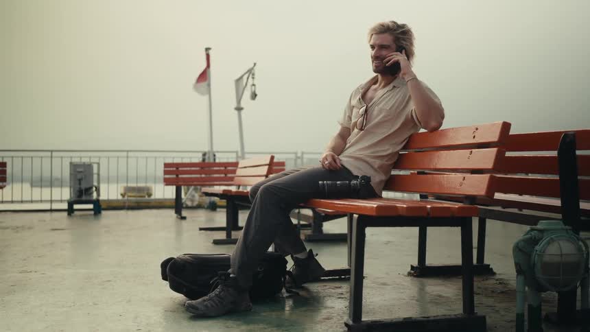 Man sitting on a bench in a park along the coast as he talks on his phone