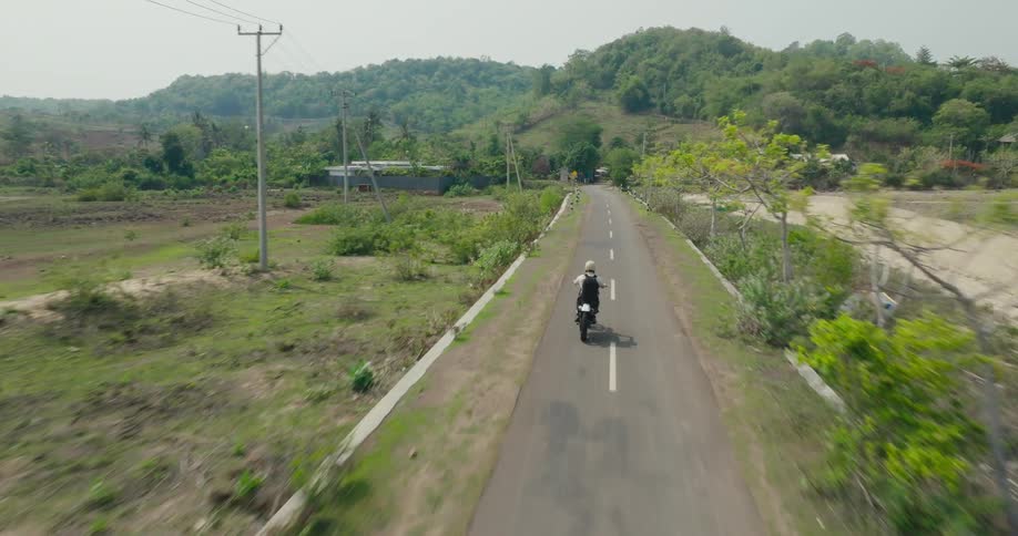 An aerial view of a motorcyclist cruising along a winding road through a lush tropical landscape