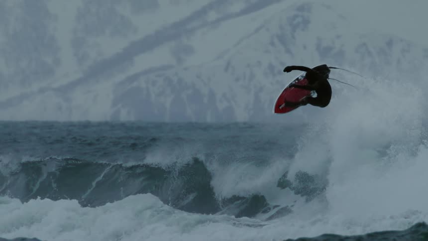 Water splashes as a surfer wearing a wetsuit leaps out of the water on his surfboard and turns with snow covered mountains in the distance