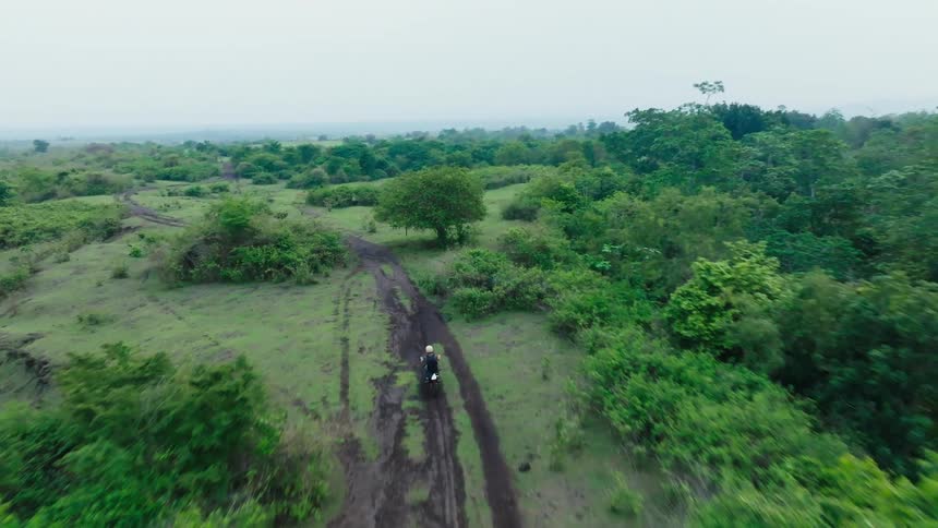 A person rides a motorcycle on a muddy path through a lush green landscape with scattered bushes and trees