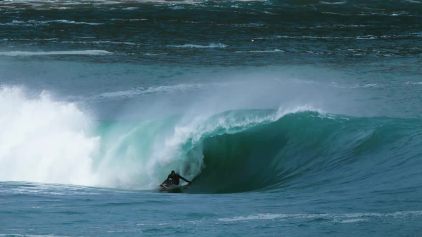 A surfer in a black wetsuit rides a large wave going through the barrel before the wave crashes