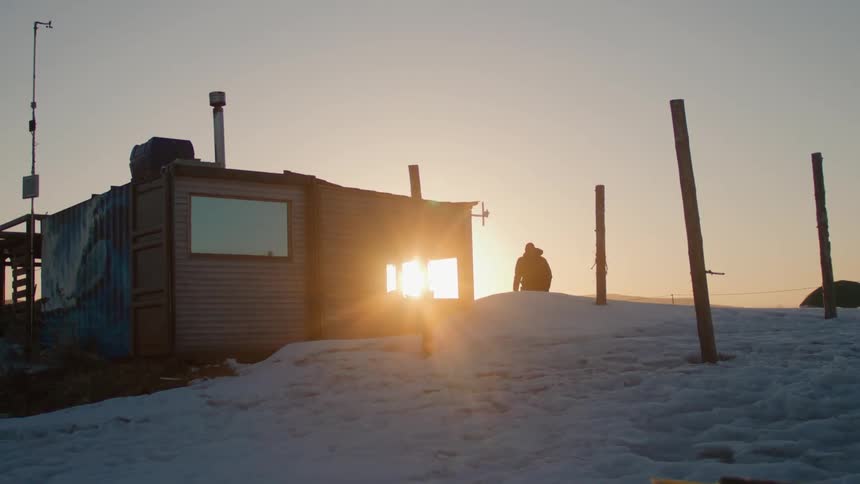 Person walking next to a cabin in the snow with the sunrise on the horizon