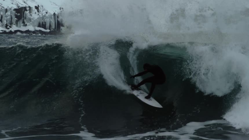 A surfer rides a powerful wave against the backdrop of a snowy mountainous coastline capturing a dramatic and adventurous moment