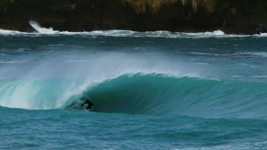 A surfer wearing a black wetsuit rides a large wave hear cliffs