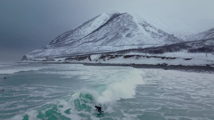 A surfer rides a wave in icy waters with a snowcovered mountain and rugged coastline in the background