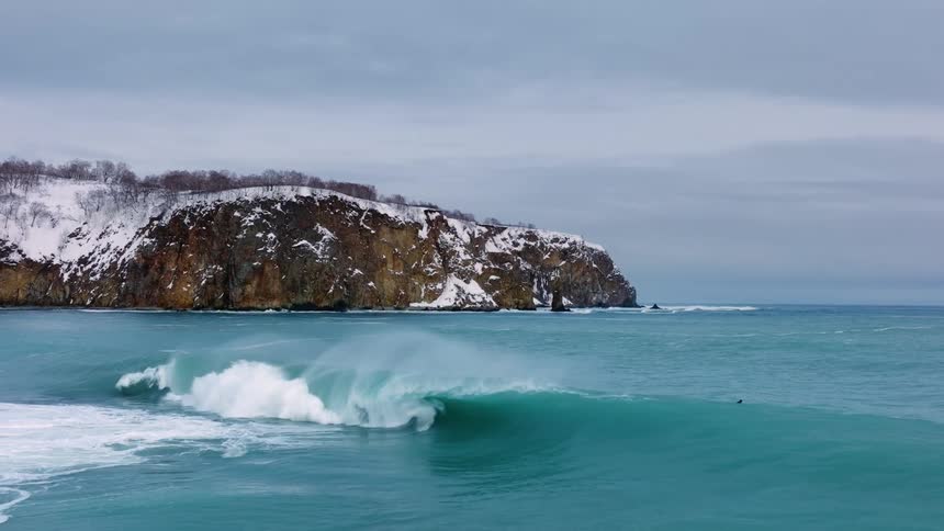 Surfers catch waves in a turquoiseblue sea with a towering snowcovered cliff in the background