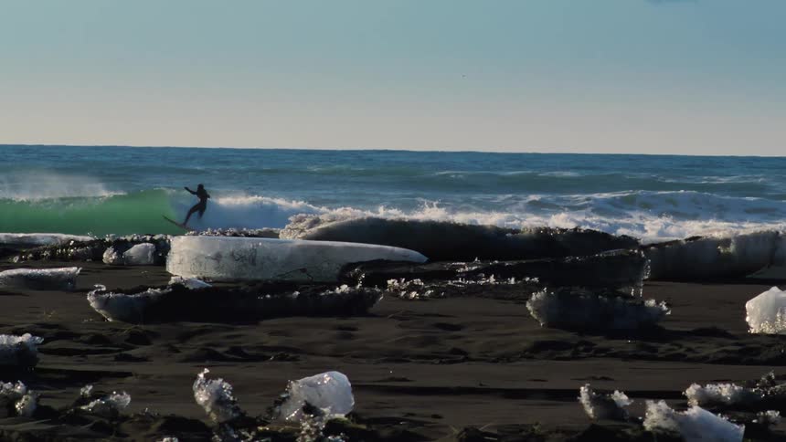 A surfer rides an ocean wave near an icy coastline with black sand beaches