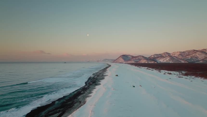 A crescent moon hangs over a pristine snow covered beach contrasting with the dark rugged mountains in the distance