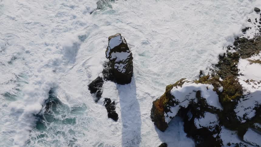 Waves roll in and splash against snow covered rock formations along the coast