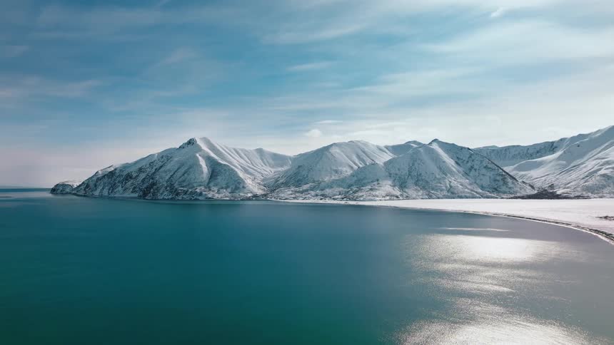 A breathtaking view of a snow capped mountain range meeting the crystal clear waters of the sea under a bright blue sky