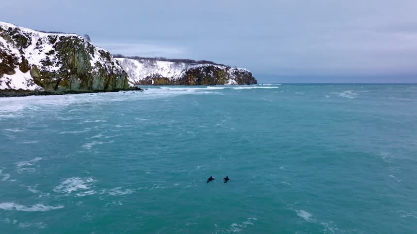Two people surfing in a large wave in a body of water with a snow covered cliff in the background