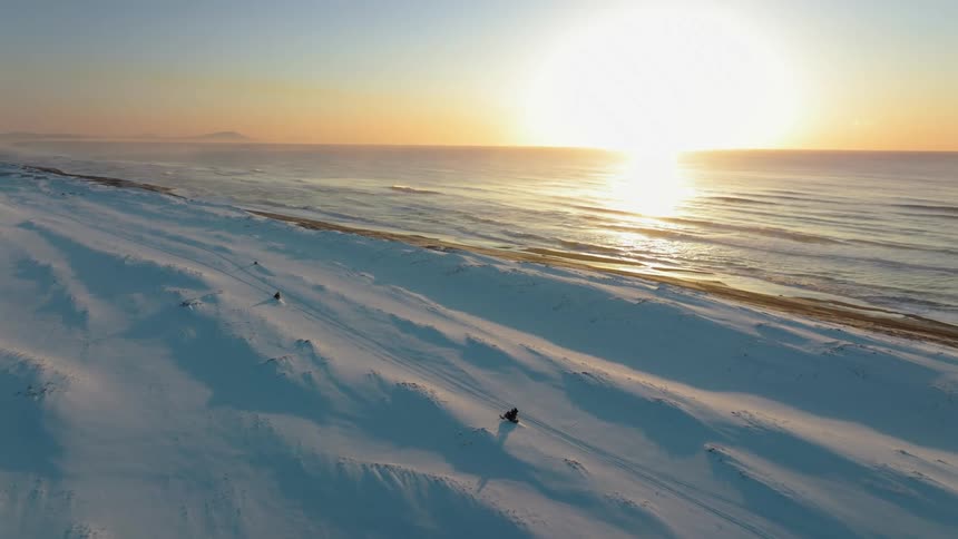Aerial view of people riding snowmobiles in a seaside setting with the sun on the horizon
