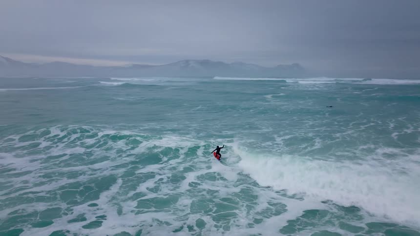 A person surfing a wave in the sea near the rocky coast in a wintery environment