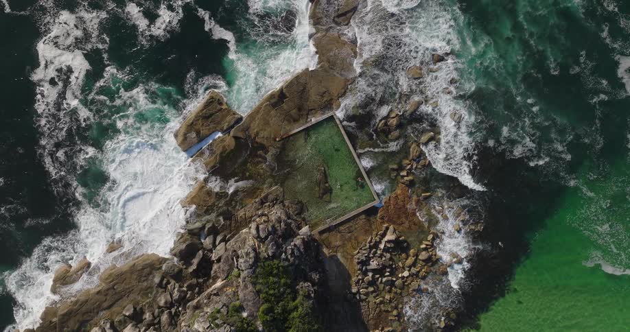 An aerial view of a rectangular rock pool carved into the coastal rocks with waves crashing around it
