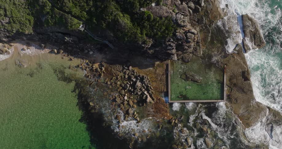 Birds eye view of the waves crashing on cliff side shoreline near a sandy cove