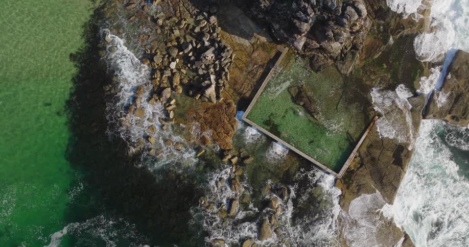 A camera looks down over water crashing on a rocky shore