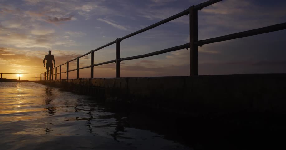 A person is walking along a pier with railings at sunset