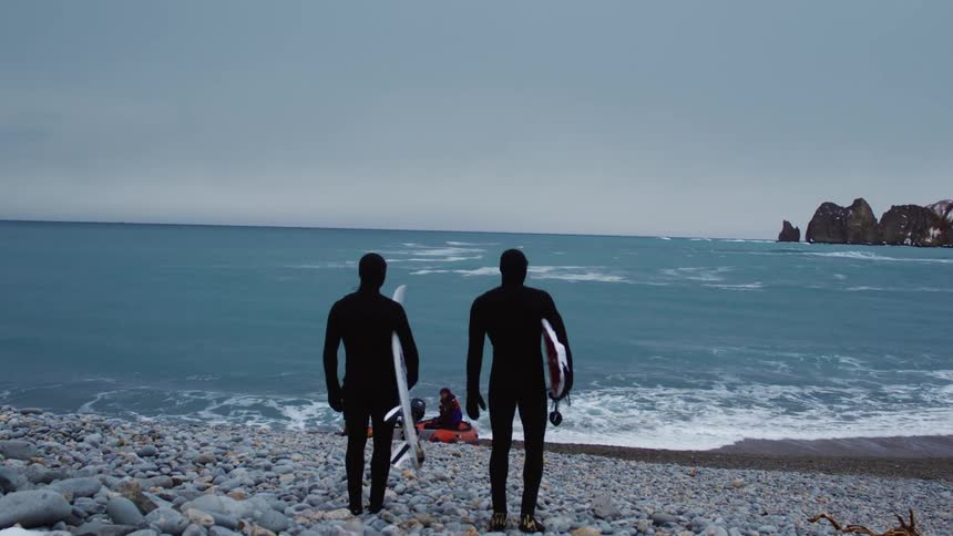 Two surfers in black wetsuits walk towards the ocean with their surfboards on a rocky beach