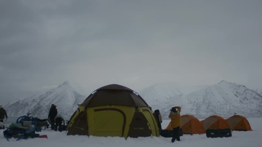 A group of people is setting up camp in a snowy landscape with a backdrop of mountains several tents already assembled on the white snow