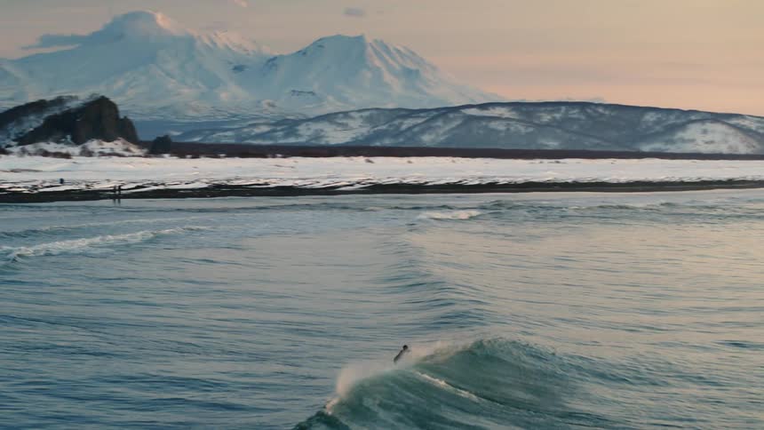 A surfer riding a wave in a cold snowy landscape at sunset