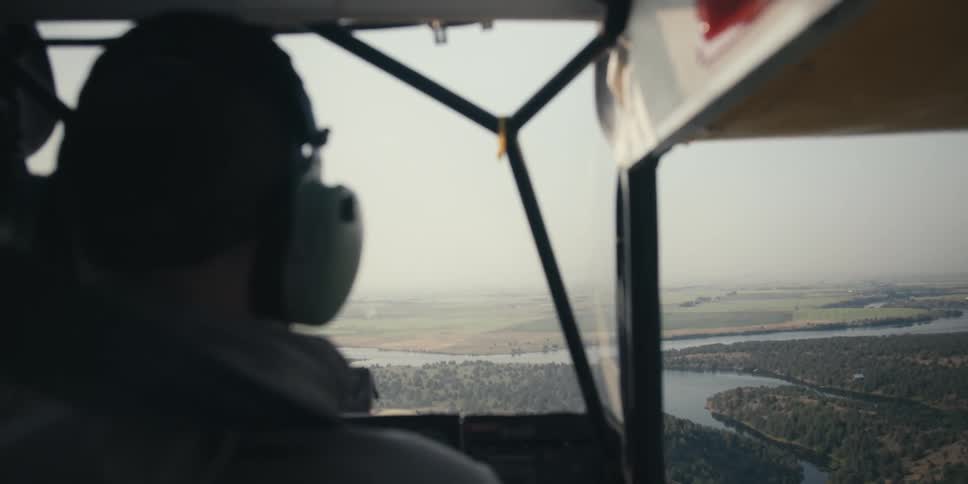 A view from inside a small aircraft with a pilot wearing headphones flying over a scenic landscape