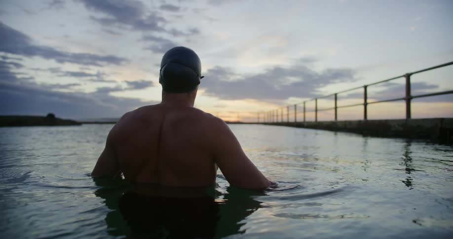 A swimmer wearing a swim cap and goggles is standing in a calm outdoor pool at dawn facing the horizon with a fence running along the right side of the pool