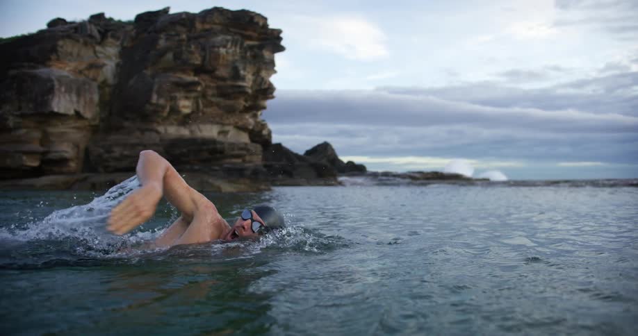 A swimmer wearing goggles and a swim cap performs a freestyle swimming in the ocean near a rocky cliff under a cloudy sky