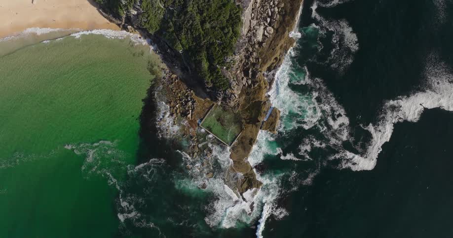 An aerial view of a natural pool on a rocky coastline surrounded by clear green water and tall waves