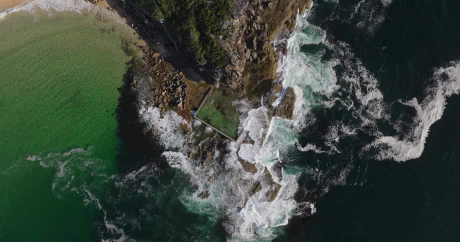 A rectangular ocean pool surrounded by rocks where waves crash on a rugged coastline with a sandy beach to the left